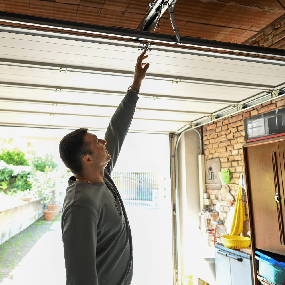 A man standing in a garage reaches up to manually close or open the overhead garage door. The garage interior includes shelves, cabinets, and gardening tools. Sunlight is visible outside.