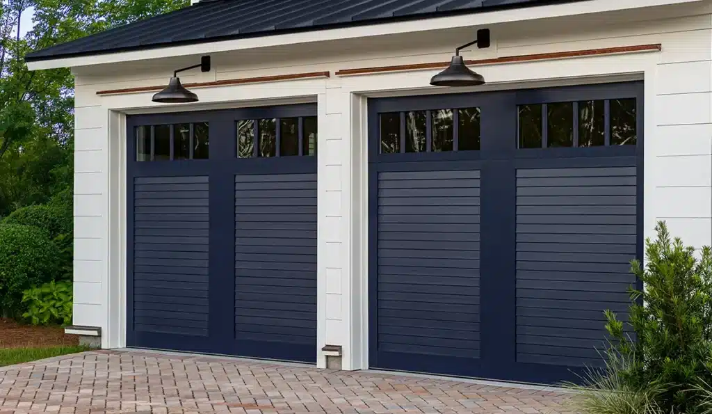 Two dark blue garage doors with horizontal panels and small upper windows on a white building, topped with black lamps, and a brick driveway in front. Shrubs and greenery surround the area.