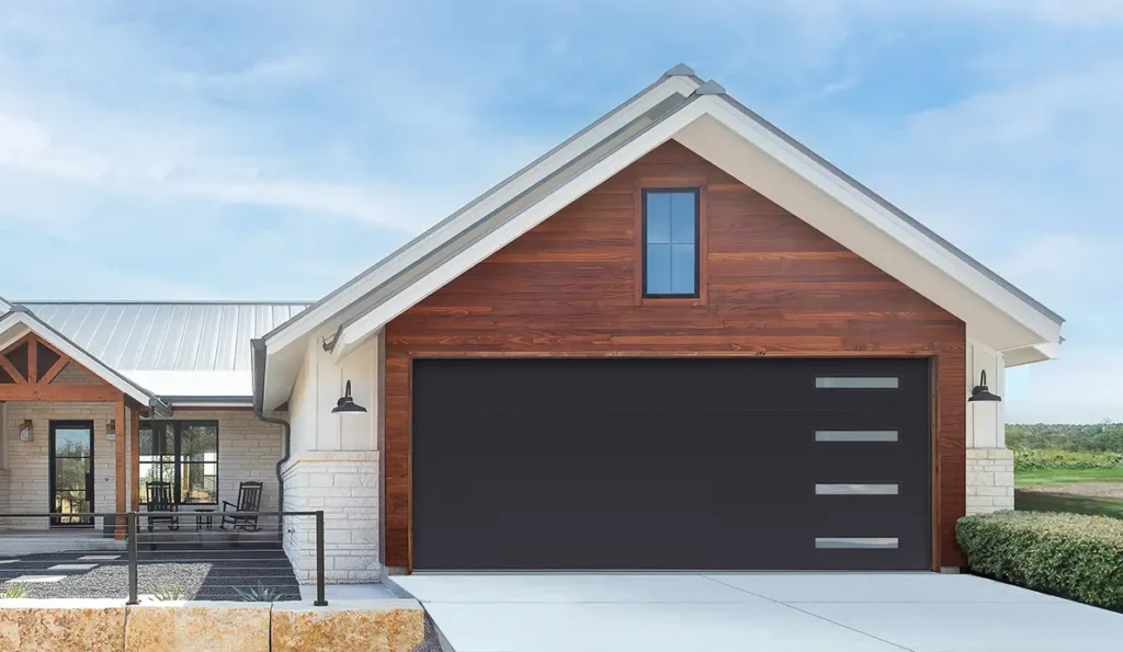 Modern house with a large dark garage door featuring three horizontal windows, wooden siding above white stone walls, a small upper window, and a landscaped front yard under a blue sky.