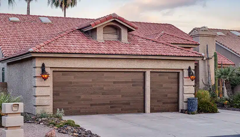 A house with a red tile roof and beige stucco walls features a two-car garage with wood-paneled doors. Small lamps are mounted on either side of the garage, and desert landscaping surrounds the driveway.